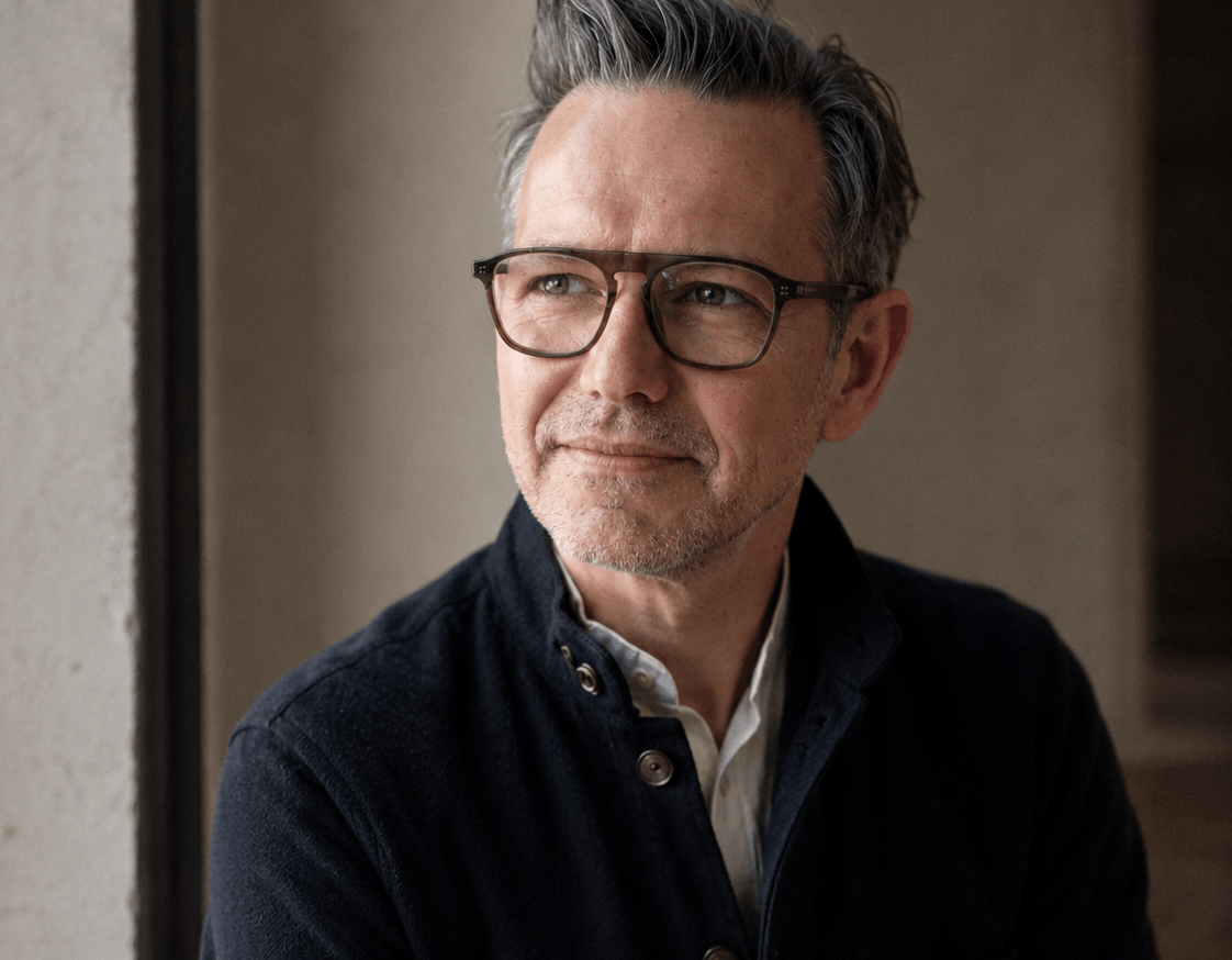 A portrait of a man with short grey hair and glasses, wearing a dark jacket over a light shirt, looking thoughtfully off-camera next to a window.