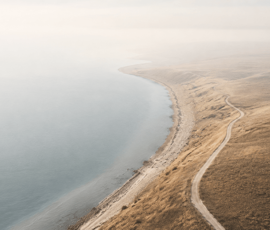 An aerial photograph of a misty coastline with a winding dirt road tracing the edge of golden, grassy hills beside a calm, pale sea.