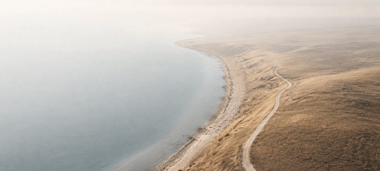 An aerial photograph of a misty coastline with a winding dirt road tracing the edge of golden, grassy hills beside a calm, pale sea.