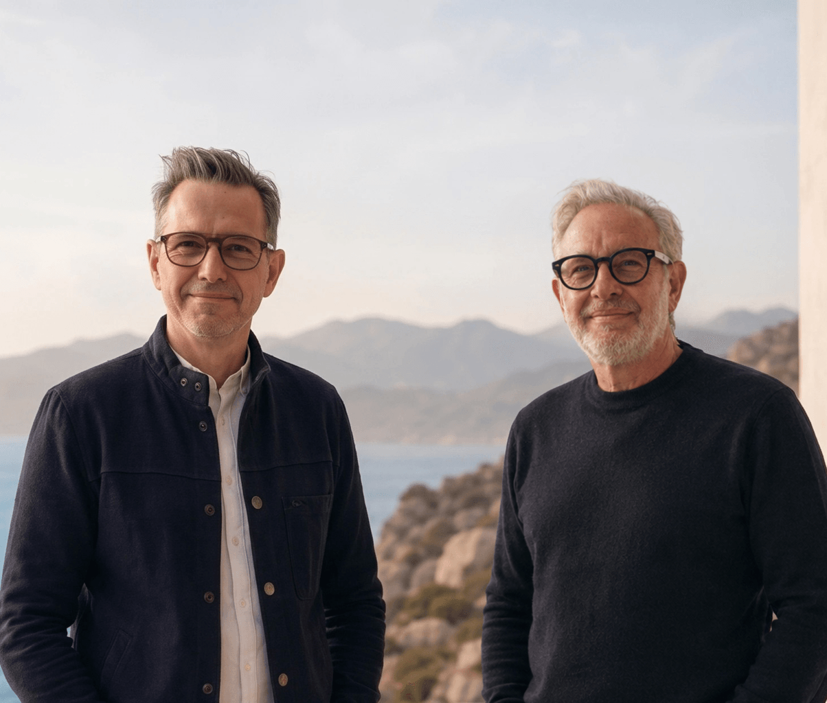 A portrait of two smiling men standing outdoors next to a building, with a scenic view of bright blue ocean water and distant mountains in the background. The man on the left wears a dark jacket over a light collared shirt and glasses, while the man on the right wears a dark crewneck sweater and thick black-rimmed glasses.