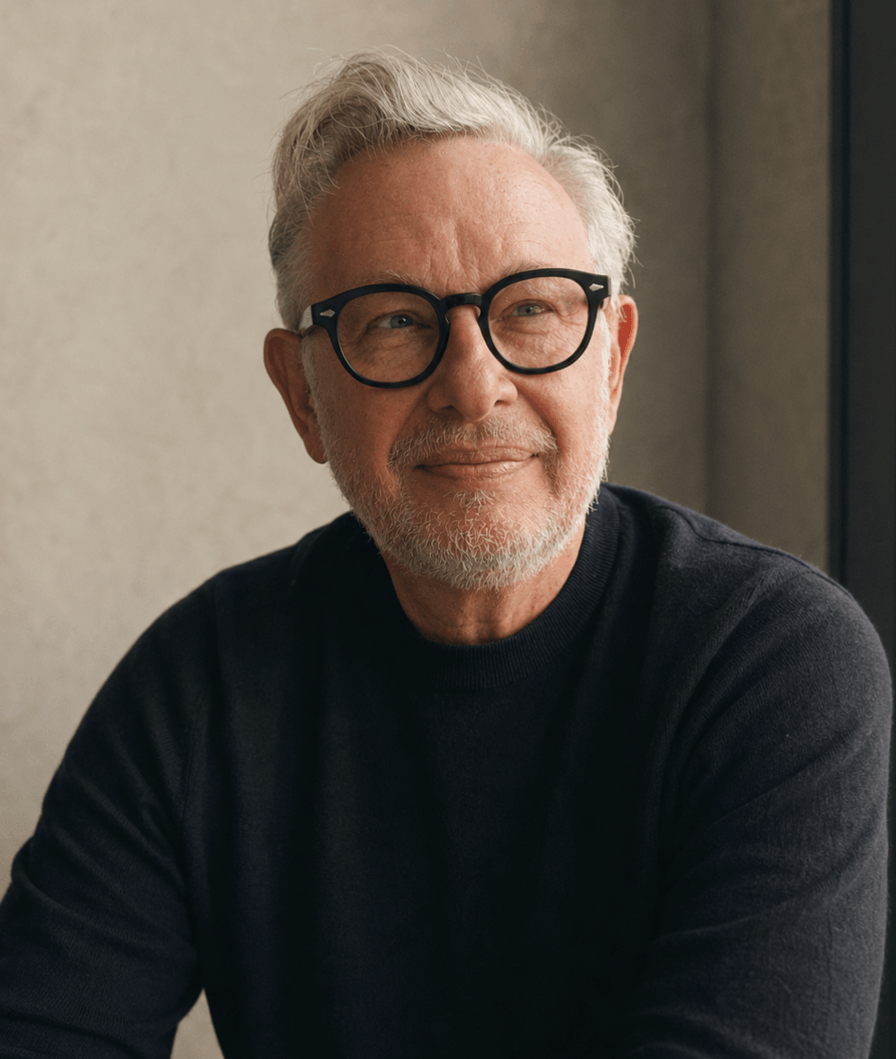 A close-up portrait of a smiling man with white hair and thick black-rimmed glasses. He is wearing a dark crewneck sweater and is softly lit against a neutral, textured wall.