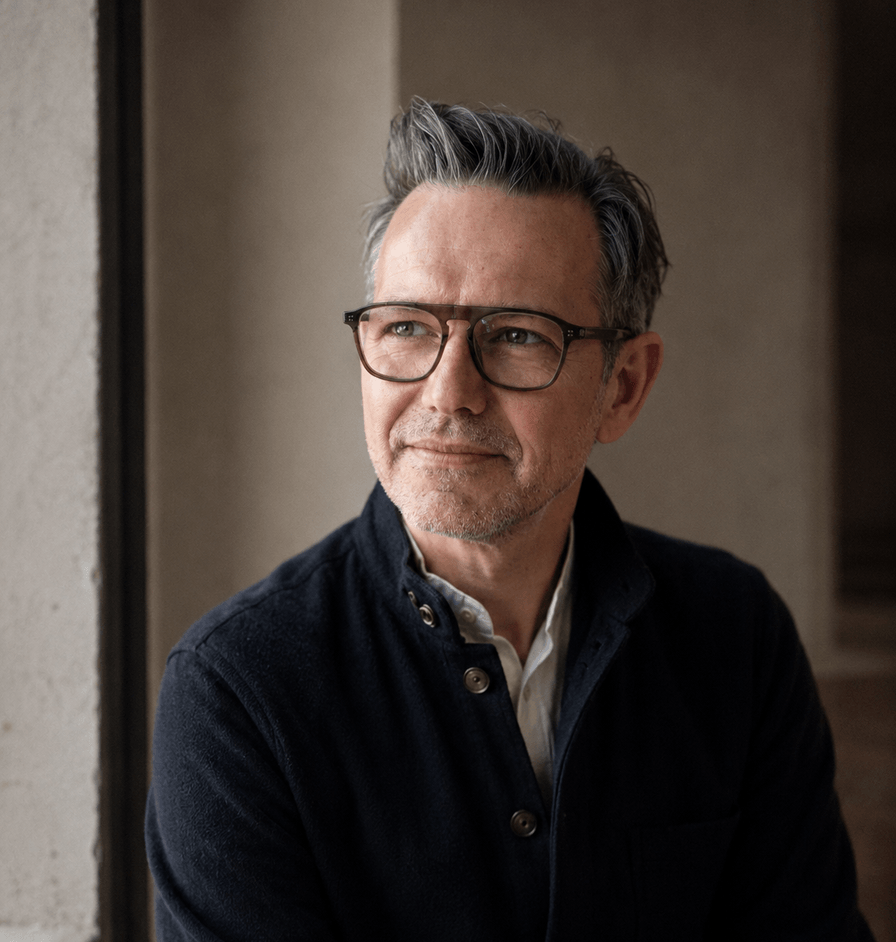 A portrait of a man with short grey hair and glasses, wearing a dark jacket over a light shirt, looking thoughtfully off-camera next to a window.