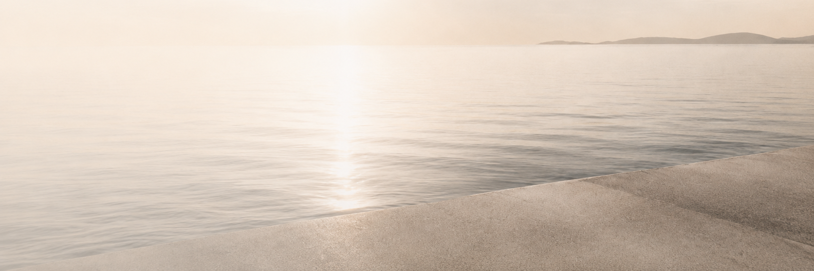 A very bright, hazy landscape looking out over calm water from a smooth concrete edge in the foreground, with a faint horizon line in the distance.