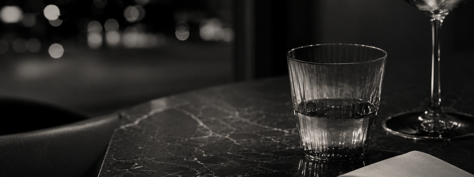 A black and white close-up of a dark marble table showing a glass of water, a folded napkin, a small dish, and the base of a wine glass, with out-of-focus city lights behind.